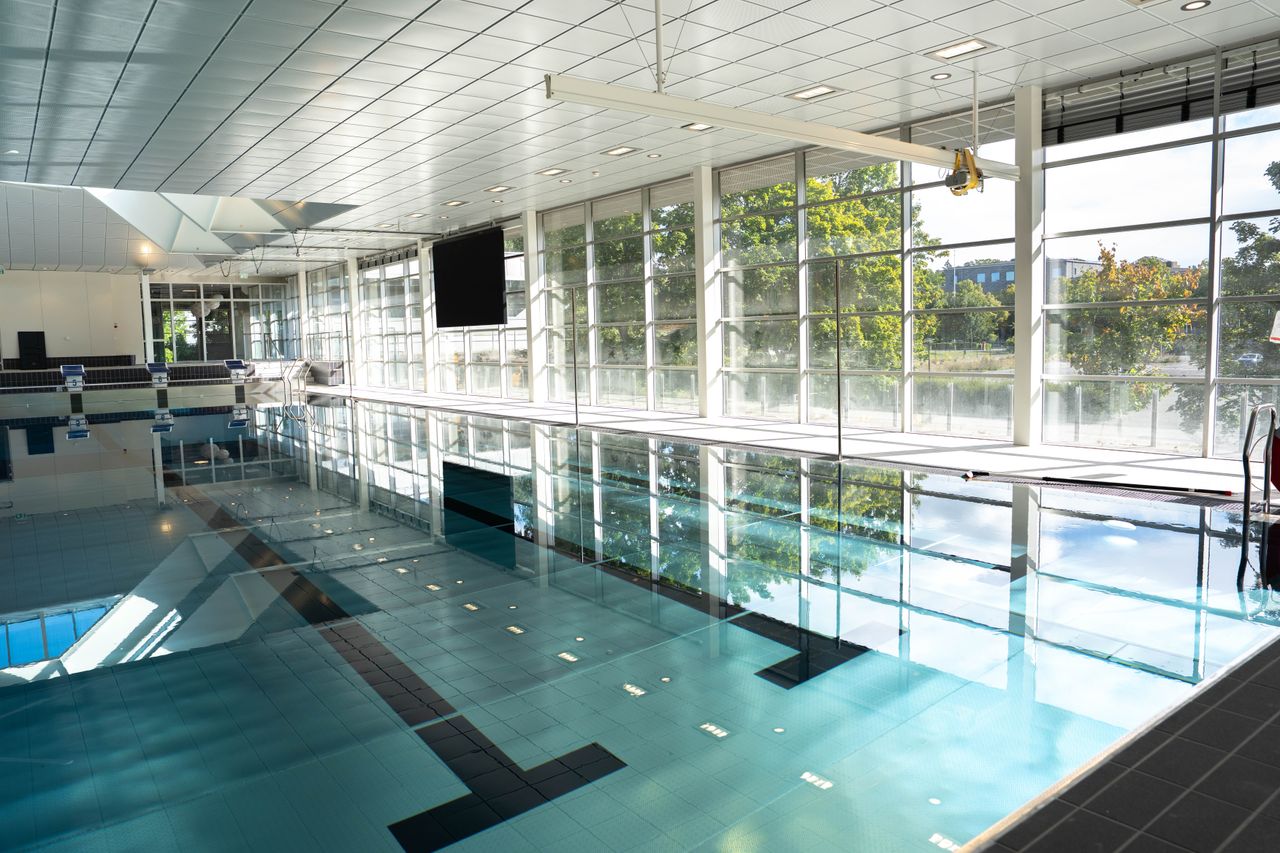 En inomhuspool med klart blått vatten, stora fönster som släpper in solljus och träd som syns utanför. Poolområdet är tomt och rent, med speglingar på vattenytan.