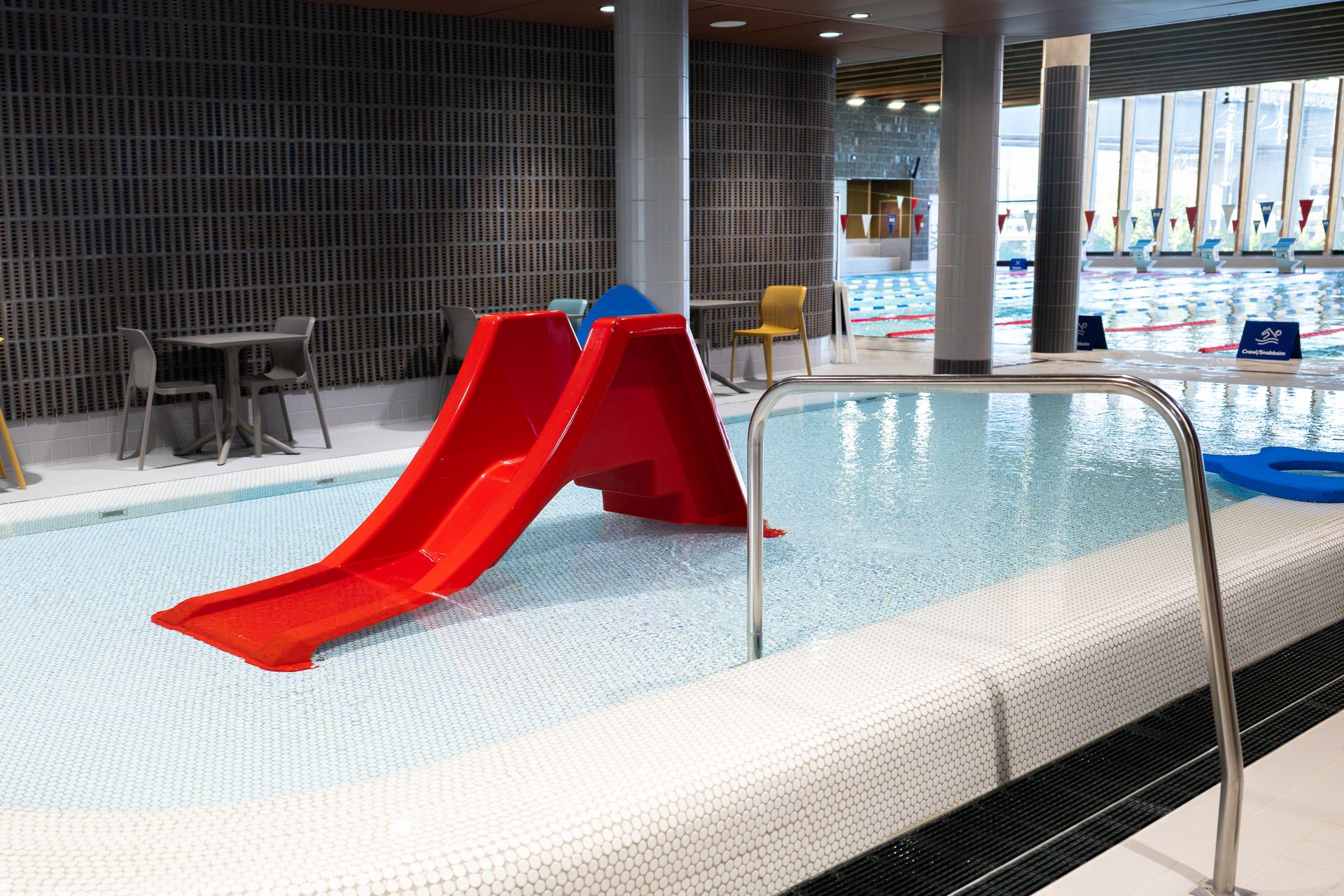 En liten inomhuspool med en röd rutschkana, bord och stolar i närheten samt en större simbassäng som syns i bakgrunden genom glasrutorna.
