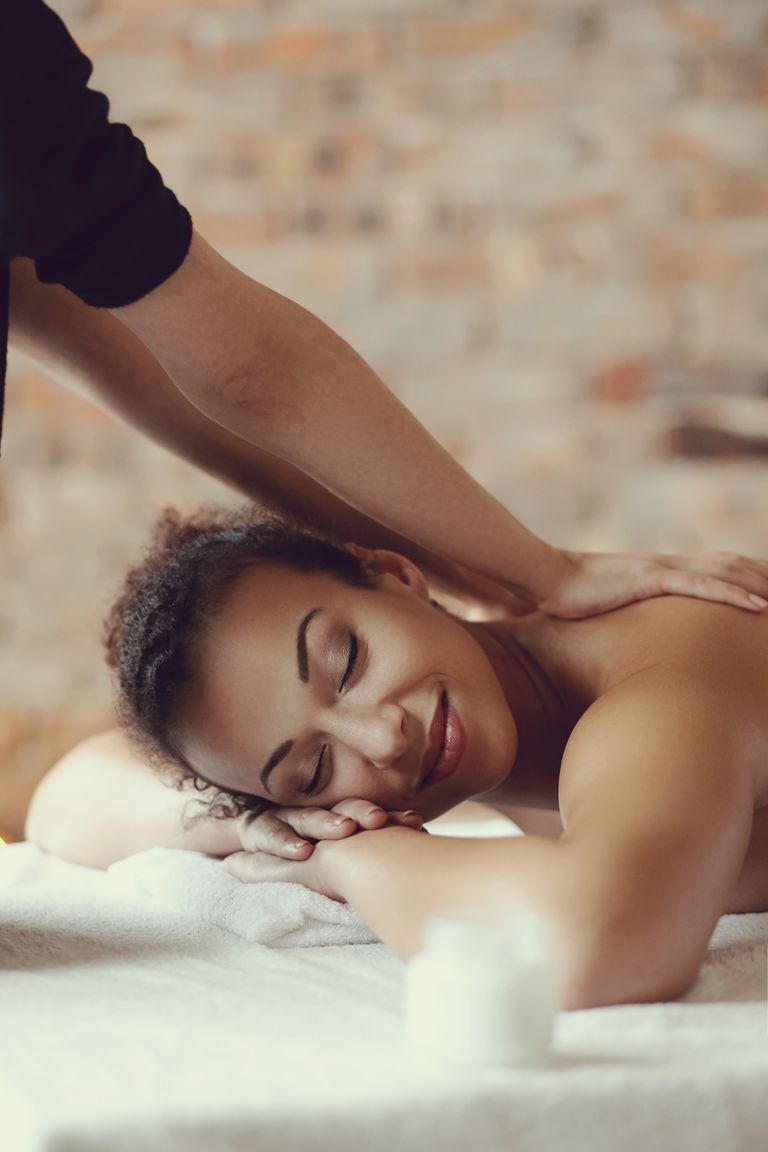 En kvinna ligger på en massagebänk med slutna ögon och ler milt medan en massör masserar hennes axlar. I förgrunden syns en suddig burk, och i bakgrunden en tegelvägg.